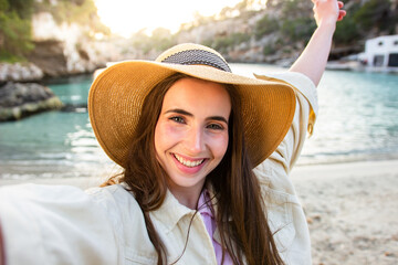 Selfie of a happy traveller with a hat in front of the beach at sunset. Beautiful woman taking a portrait.