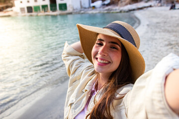 Selfie of a smiling traveller with a hat in front of the mediterranean sea at sunset. Beautiful woman taking a portrait in front of the ocean.