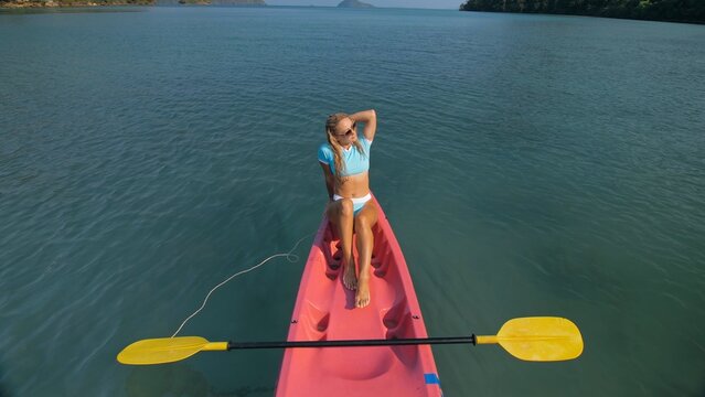 Pretty slim woman in elegant swimsuit lies in pink plastic kayak drifting on azure ocean at exotic resort upper view. Traveling to tropical countries. Girl is sailing on kayak in ocean, view above.