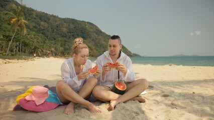 Fototapeta premium The cheerful love couple holding and eating slices of watermelon on tropical sand beach sea. Romantic lovers two people caucasian spend summer weekend in outdoor. Hat, backpack white shirt beachwear.