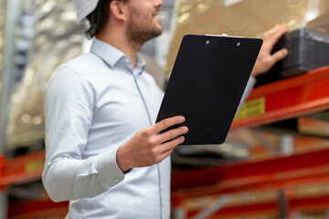 logistic business, shipment and people concept - close up of businessman in helmet with clipboard checking goods at warehouse