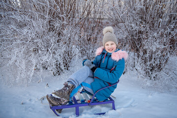 In winter, a child puts on jackets, trousers, a hat and boots in winter on white snow on the street and in a park in nature, sledding and playing winter fun. Active walks in winter for children