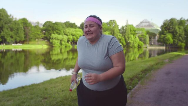 Tired fat woman in sportswear running in summer park. Realtime