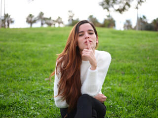 A young woman in the park sitting on the grass sensually asking for silence.