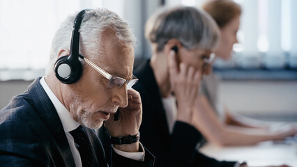 Mature businessman in headset talking near blurred colleagues in office