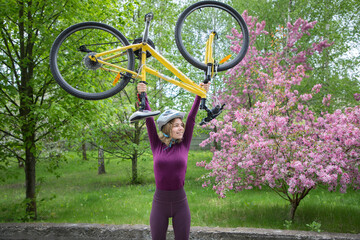 strong slender cheerful young female cyclist in a helmet lifting her modern bike over her head....