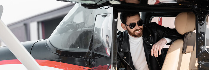 bearded pilot in stylish sunglasses and leather jacket smiling while sitting in cockpit of...
