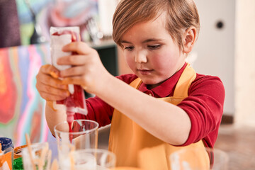 Boy pouring red paints into the glass while standing at the table and drawing with inspiration