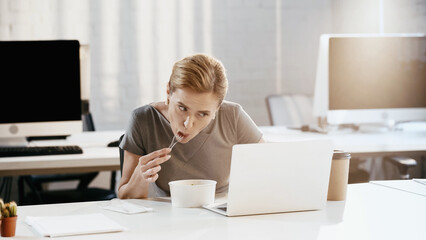 Businesswoman eating fresh salad near laptop and papers in office