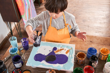 Cute little boy pouring paints into the canvas while painting diligently at the table at art school