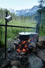 Food is prepared in the forest camping. In the foreground iron mug and two pans hang on the cooking rack on a campfire, in the background a valley, trees and a snowy peak of a rocky mountain.