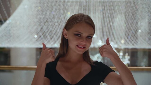 Caucasian girl gestures cool sign. A beautiful European woman in a black dress on a background of a large crystal chandelier. Portrait of a pretty lady.