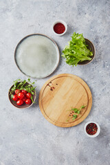 Wooden round cutting board, greens, vegetables and seasonings on light background