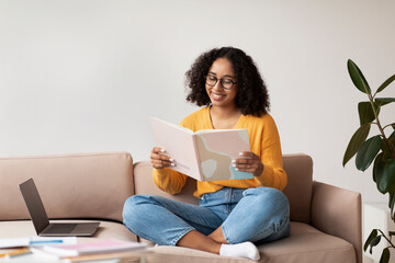 Cheery African American woman sitting on couch with laptop and book, doing home assignment, reading at home, free space