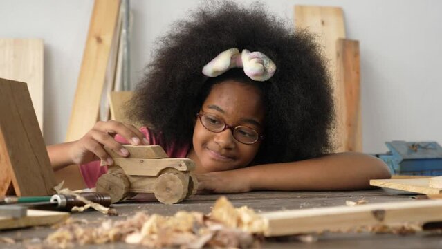 A cheerful dark skin African teen girl wearing eyeglasses and afro-hair style, playing, moving a wooden toy car along the table, smiling, being happy, and relaxed. Carpentry and craftmanship concept.