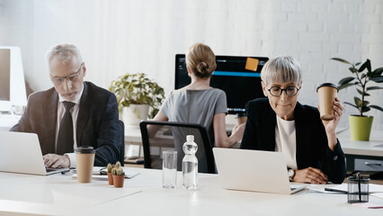 Businesswoman holding paper cup near laptop, papers and colleague in office