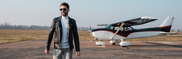 happy pilot in stylish leather jacket and sunglasses walking near helicopter, banner.