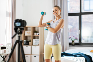 Fototapeta na wymiar fitness, sport and video blogging concept - happy smiling teenage girl or blogger with camera on tripod and dumbbells recording online class at home