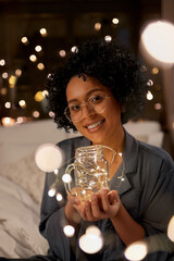 people, bedtime and rest concept - happy smiling woman in pajamas and glasses sitting in bed with garland lights in glass mug at night