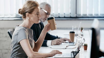 Side view of businesswoman holding paper cup and using laptop in office
