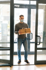 Vertical portrait of courier male in medical face mask holding boxes hot pizza and payment POS terminal entering lobby of apartment building. Delivery man walks into office building to meet customer.