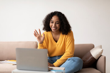 Happy African American lady video calling via laptop, waving hello at computer webcam in living room