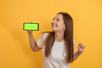 Naklejka premium Portrait of a joyful girl with a phone with a green screen in her hands, cheerful, happy, on a yellow background