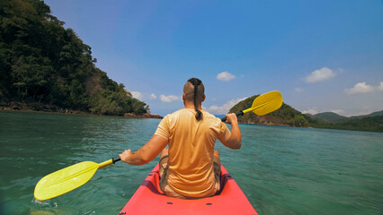 Strong man with thin plait hairstyle rows pink plastic kayak putting up paddle on sea against hills with wild jungles and blue sky. Young guy is sailing on kayak in ocean. Traveling to tropics.