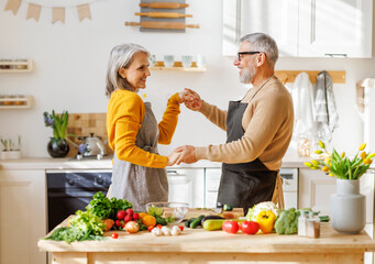 Happy senior elderly couple husband and wife embracing and dancing while cooking together in kitchen