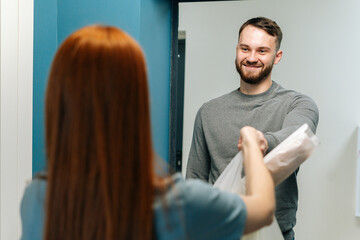 Fototapeta premium Cheerful bearded courier male delivering groceries bag with to unrecognizable female housewife customer on doorway at home. Rear view of young woman client receiving takeout food meal from deliveryman
