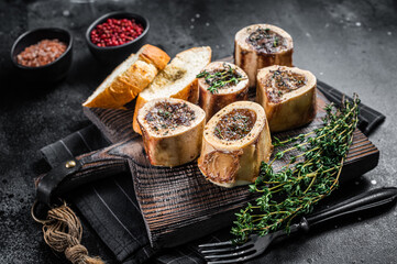 Roast marrow calf bones on wooden board with bread and herbs. Black background. Top view