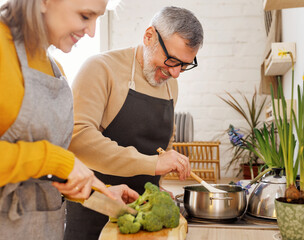 Happy joyful senior couple in aprons preparing vegetarian soup together in cozy kitchen