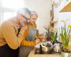 Happy joyful senior couple in aprons preparing vegetarian soup together in cozy kitchen