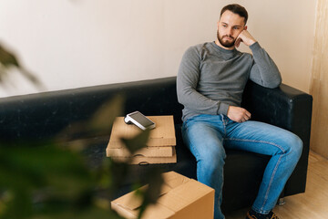 Bored courier male waiting customer while sitting on sofa with boxes pizza, cardboard box and payment POS wireless terminal in hall of apartment or office building. Concept of online shopping.
