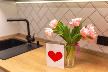 Bouquet of pink tulips in a glass vase with a postcard in the white interior of the kitchen