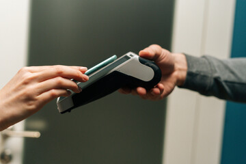 Close-up side view of unrecognizable delivery man giving POS wireless terminal to making contactless payment via smartphone card to young female customer, on entryway at apartment, cropped shot.
