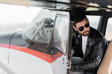 bearded pilot in sunglasses and leather jacket opening door of helicopter and looking away.