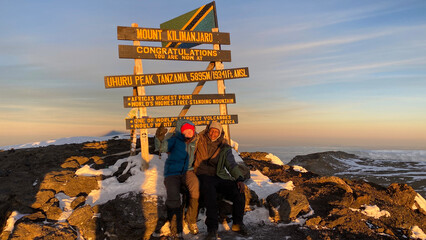 A happy hiker sits on top of Mount Kilimanjaro with his black guide. Breathtaking sunrise in the...