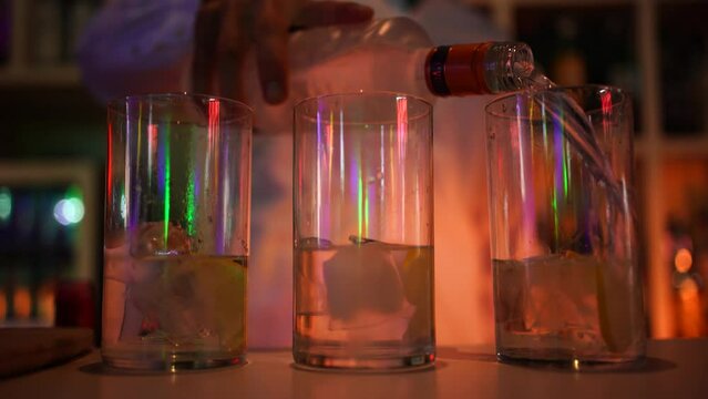 Camarero sirviendo alcohol detr&aacute;s de una barra de discoteca con luces de ne&oacute;n. Los vasos est&aacute;n llenos de hielo y contienen fruta, la acci&oacute;n es servir una copa.