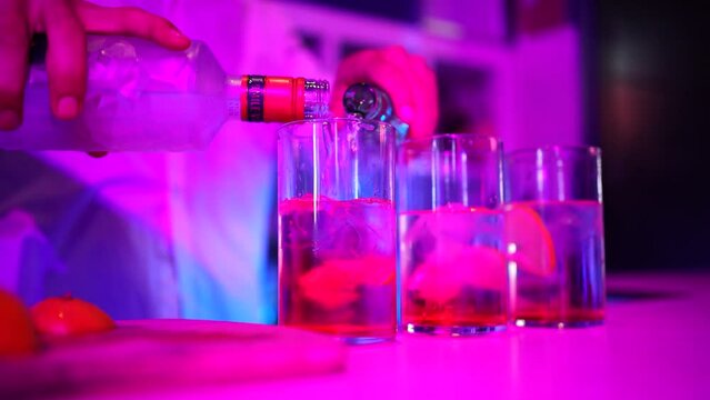 Se sirve una copa de ginebra con un refresco en la barra de una discoteca con luces de ne&oacute;n. El camarero llevo camisa blanca, se aprecian las manos con las mangas. Se ven 3 vasos con hielos y lim&oacute;n. 