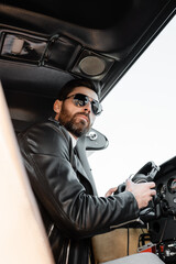 low angle view of bearded pilot in sunglasses and leather jacket holding headphones in cockpit of helicopter.