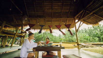 Fototapeta na wymiar The happy family eat and drink tasty beverages spending time in local floating cafe on water. Mother and daughter having breakfast outdoors. Woman and a child in a cafe. Tropical view