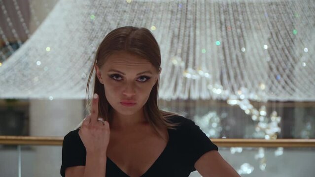 Caucasian girl gestures fuck sign. A beautiful European woman in a black dress on a background of a large crystal chandelier shows her middle finger. Portrait of a cruel lady.