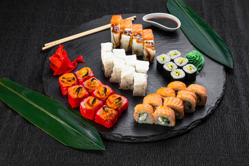 sushi rolls laid out on a dark background decorated with bamboo leaves and chopsticks