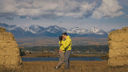 Man and woman in yellow green sportswear. Lovely couple of travelers hug and kiss near old stone enjoying highland landscape. Two travelers are walking against the backdrop of snow-capped mountains.