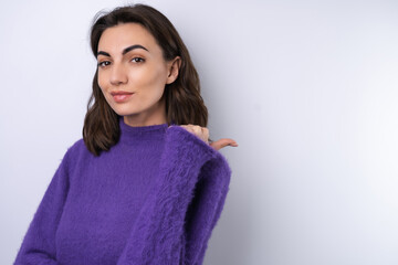 Young woman in a purple soft cozy sweater on a background of cute smiling cheerfully, in high spirits, points a finger to the right to an empty space.