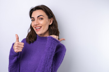 Young woman in a purple soft cozy sweater on a background of cute smiling cheerfully, in high spirits, points a finger to the right to an empty space.