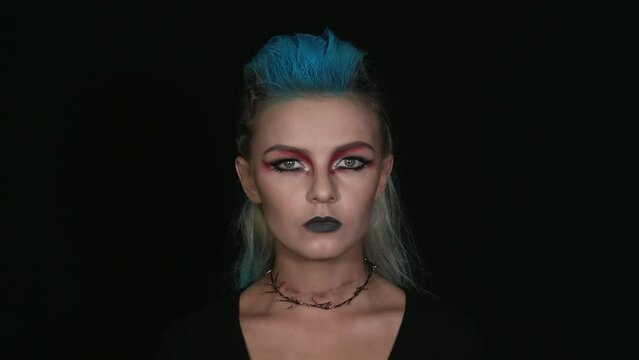 Caucasian girl looks at the camera. An informal girl with blue hair and an evil look. European woman with unusual make-up. Portrait shooting on a black background. unusual appearance