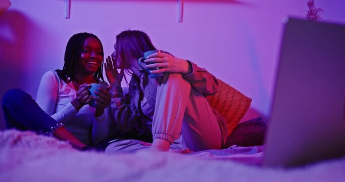 Young diverse girls gossiping in room with neon light. Low angle of smiling multiracial teen girls spreading rumors and whispering while talking to each other and sitting on bed in dark room with neon