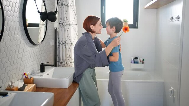 Little boy congratulates mother and gives her flower in bathroom at home.